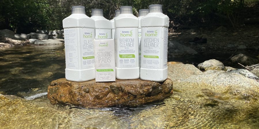 Amway Home cleaners — glass cleaner, bathroom cleaner, kitchen cleaner and scouring pads — set on a rock in a creek